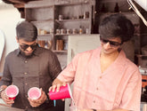 Two individuals standing behind a counter, wearing casual linen clothing; one in a pink shirt and the other in a brown shirt. The setting appears to be a casual, possibly beach-side cafe or bar.